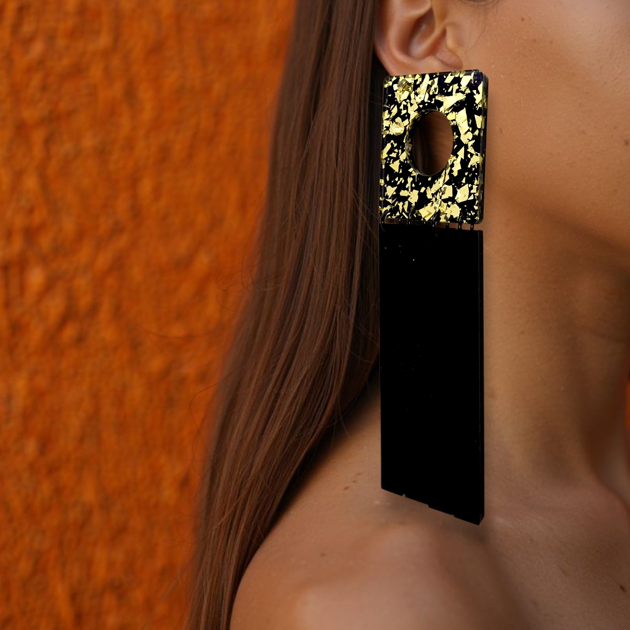 Brinco maxi geométrico preto e dourado com base quadrada marmorizada usado na orelha de uma mulher