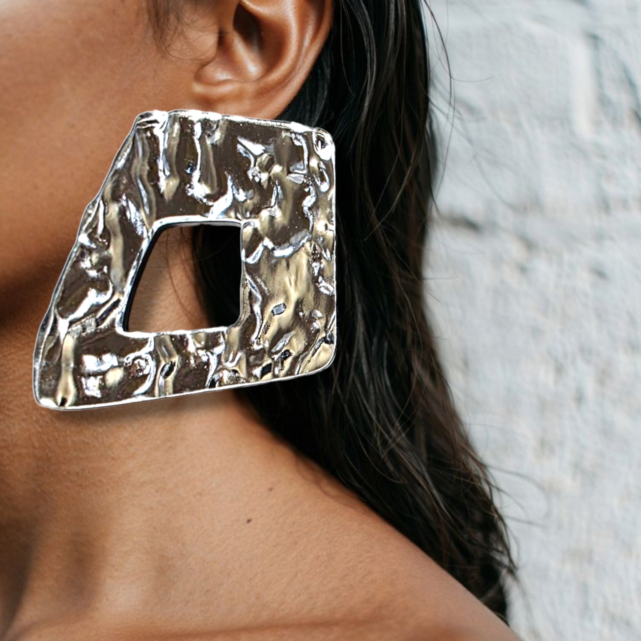 Brinco maxi prateado com textura martelada e recorte geométrico usado na orelha de uma mulher