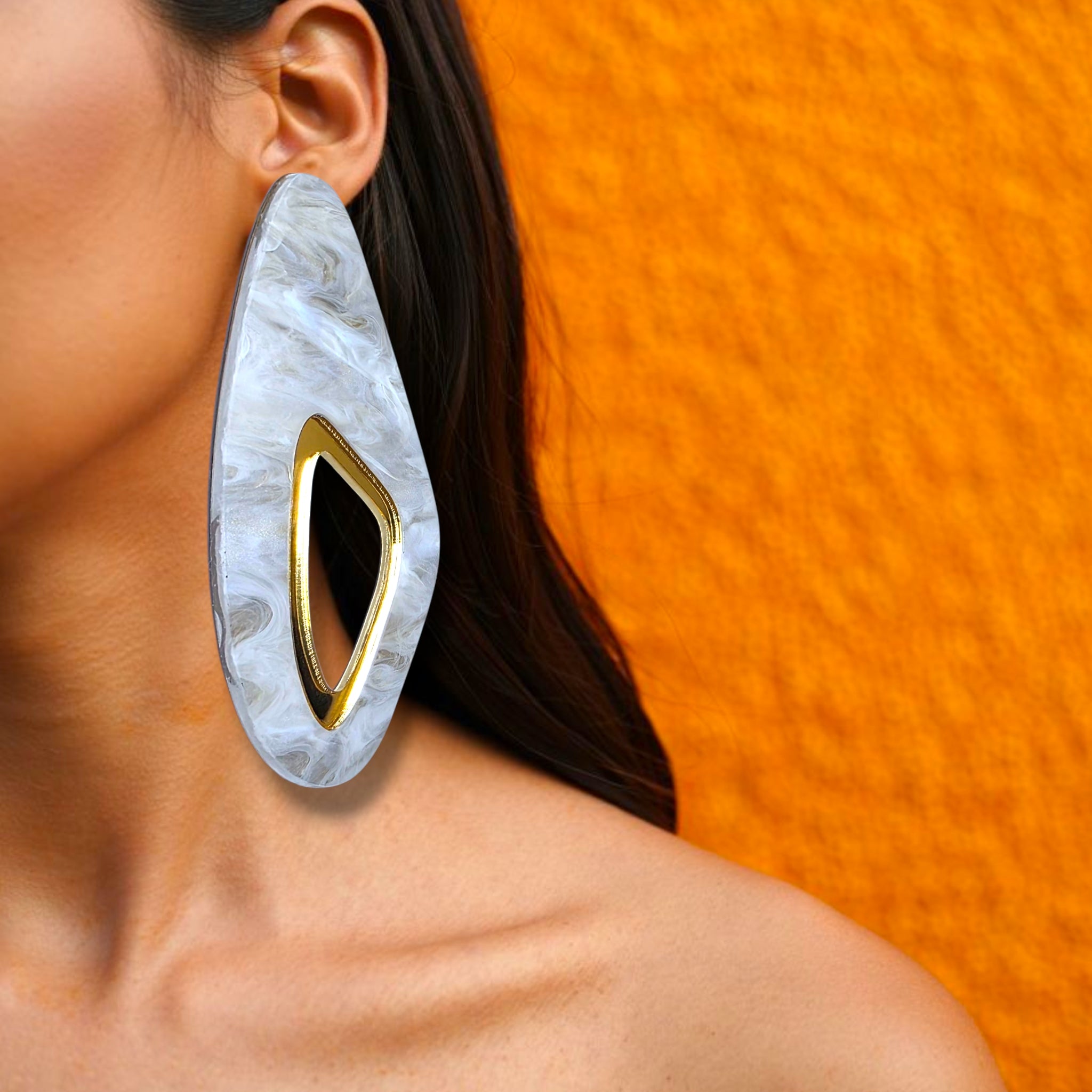 Brinco grande alongado com centro vazado dourado e base marmorizada em branco usado na orelha de uma mulher