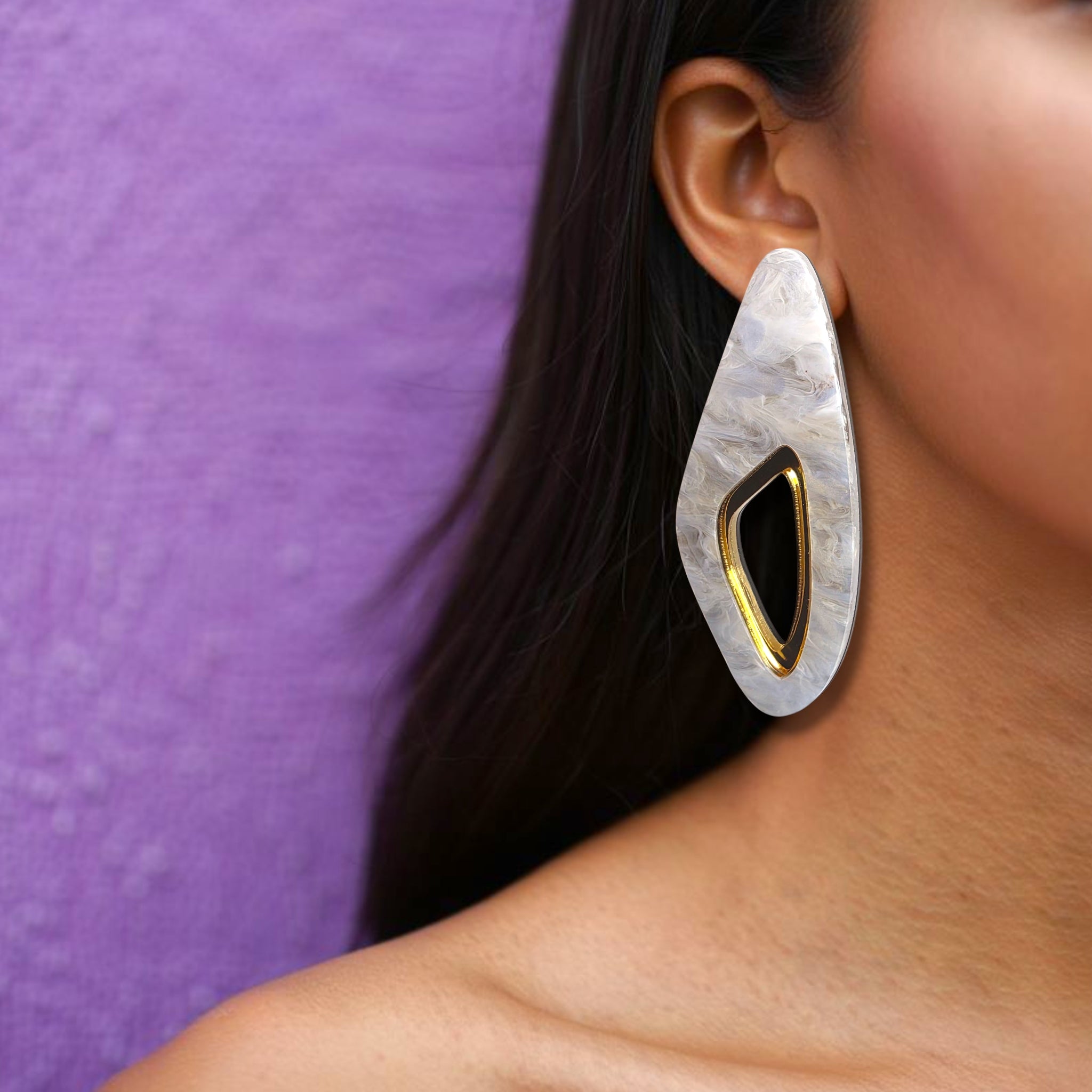 Brinco médio alongado com centro vazado dourado e base marmorizada em branco usado na orelha de uma mulher